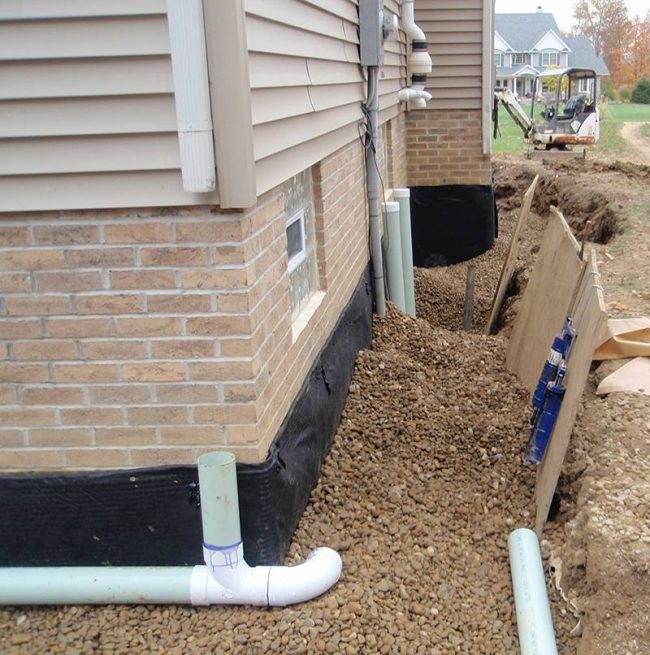 Sitework Developing Stands by Residential Waterproofing Systems with Lifetime Warranty corner ground edge of home shows gravel and drainage pipes portion of residential waterproofing system by Sitework Developing Inc in Chagrin Falls Ohio
